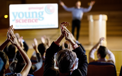 Eine klatschende Menge, im Hintergrund das Young-Science-Gütesiegel-Logo auf einer Leinwand