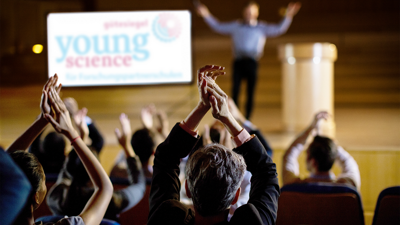 Eine klatschende Menge, im Hintergrund das Young-Science-Gütesiegel-Logo auf einer Leinwand