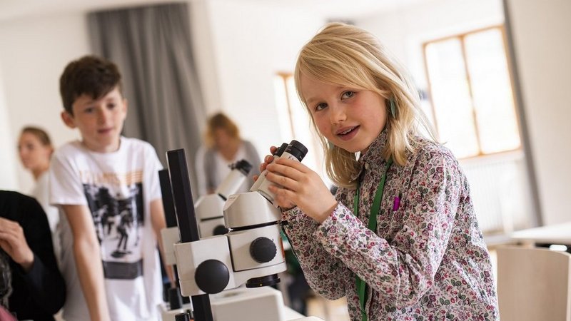 Ein Mädchen sieht von einem Mikroskop auf in die Kamera. Im Hintergrund sieht ein Bub zu. 