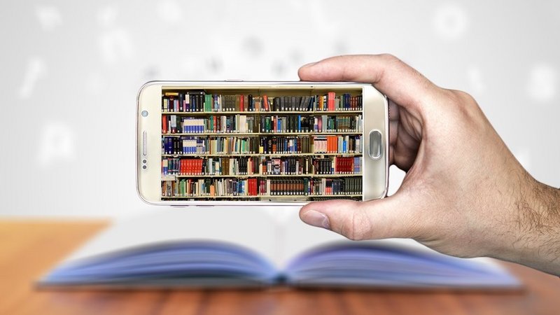 Auf einem Smartphone ist ein Bücherregal zu sehen, im Hintergrund ein aufgeschlagenes Buch