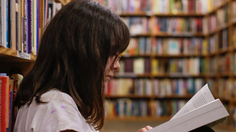 Mädchen liest an Bücherregal gelehnt in einem Buch 