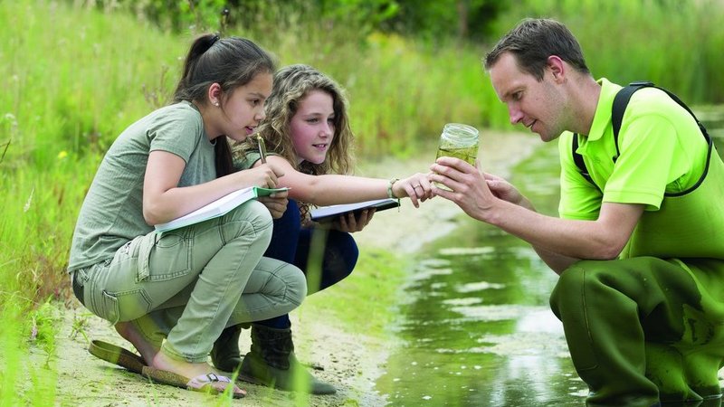 Zwei Schülerinnen und ein Forscher entnehmen eine Wasserprobe